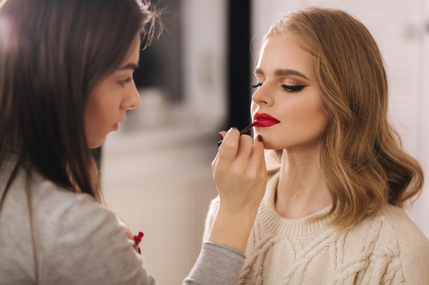 Makeup artist applies red lipstick Hand of makeup master painting lips of young beauty blond hair model Make up in process Closeup view