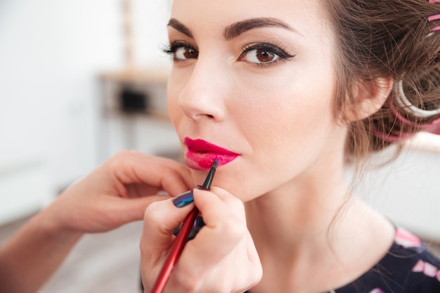 Makeup artist applying bright pink lipstick to lips of young woman in curlers