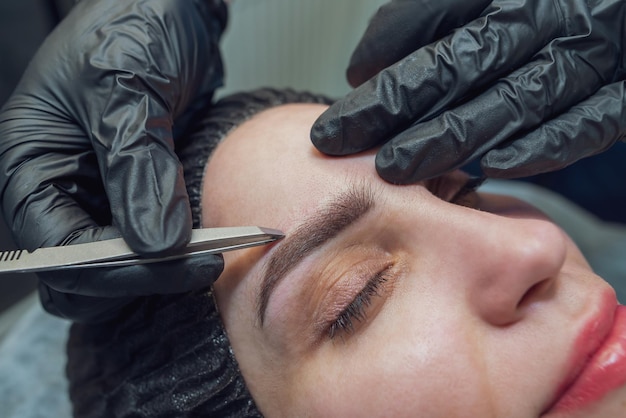 A makeup artist pinches the eyebrows of a woman with tweezers before the permanent eyebrow procedure