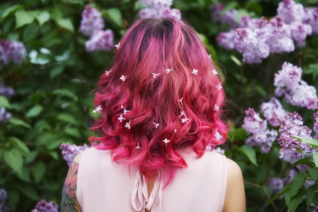 明るい赤ピンクの女の子の髪に薄紫色の花を付けたメイクヘアー。明るい飽和色の髪。ライラックの枝でポーズをとる女性