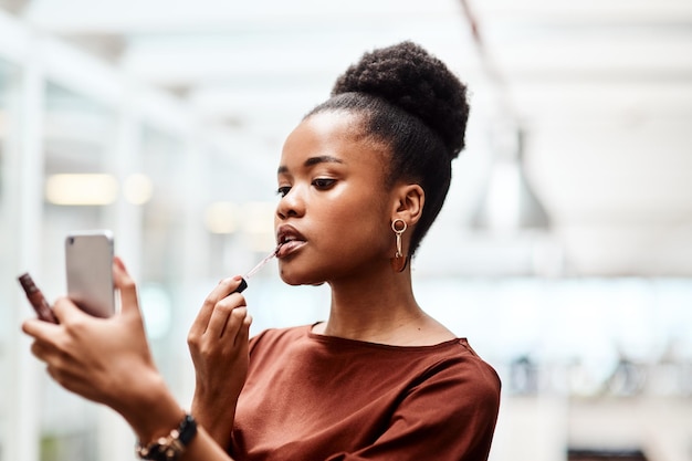 Makeup makes me feel boss Shot of a young businesswoman applying lipstick while using her cellphone as a mirror in an office