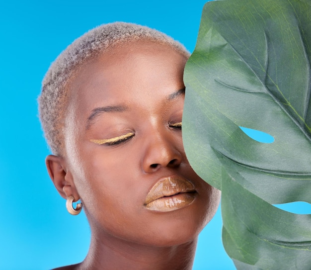 Makeup plant and a black woman on a blue background for a wellness aesthetic face beauty and an african girl or model with a leaf and organic cosmetics isolated on a studio backdrop for health