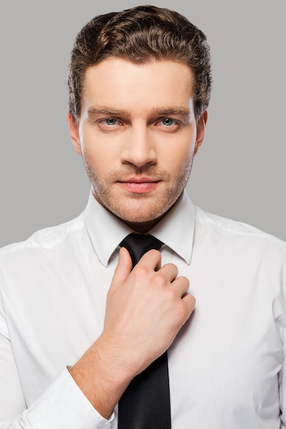 Making business look good. confident young man in shirt and tie adjusting his necktie and looking at camera while standing against grey background