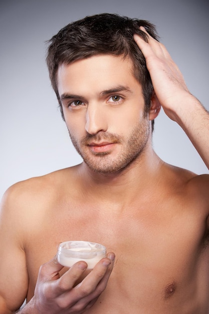 Making a charming style. Handsome young shirtless man touching his hair with hand while holding a container with gel and isolated on grey background