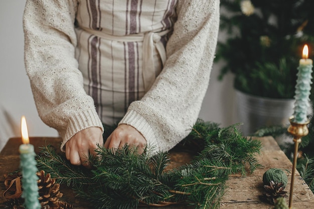 クリスマスの素朴な花輪を作る女性手モミの枝を押し、木製のテーブルに花輪を作る