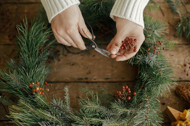 素朴な木の上のハサミで赤い果実の枝を切るセーターの女性のクリスマス リースを作る