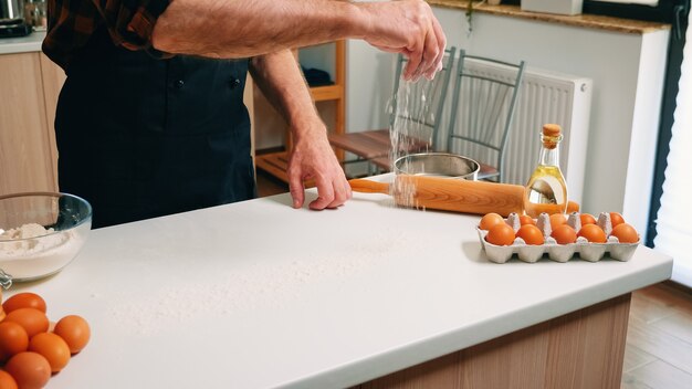 Making dough by man hands at home in kitchen using wheat flour. Retired senior baker with bonete and apron sprinkling, sifting, spreading rew ingredients baking homemade pizza and bread.