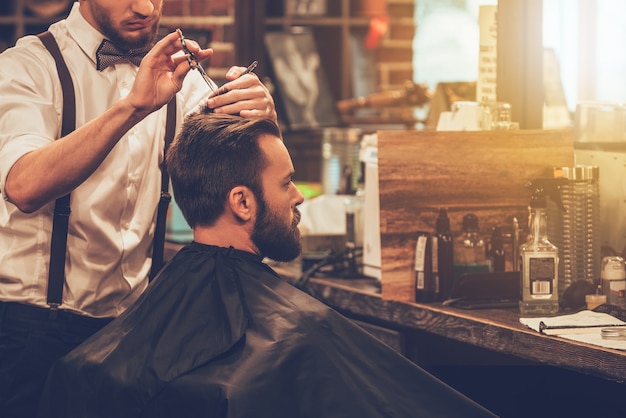 Rendere magici i capelli. giovane uomo barbuto che si fa tagliare i capelli dal parrucchiere mentre è seduto in poltrona al barbiere