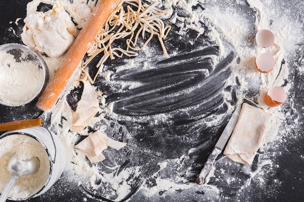 Making homemade noodles recipe concept, sprinkled wheat flour, eggs and raw pasta. Top view on blackboard or table. Cooking dough