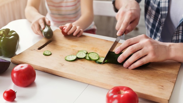 Making pleasant surprise for mom: pretty little girl assisting her dad, cutting cucumber for salad, copy space