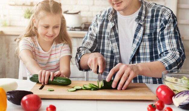 Making pleasant surprise for mom: pretty little girl assisting her dad, cutting cucumber for salad, copy space