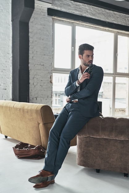 Making right decision. Full length of thoughtful young man in full suit looking away while leaning on the sofa