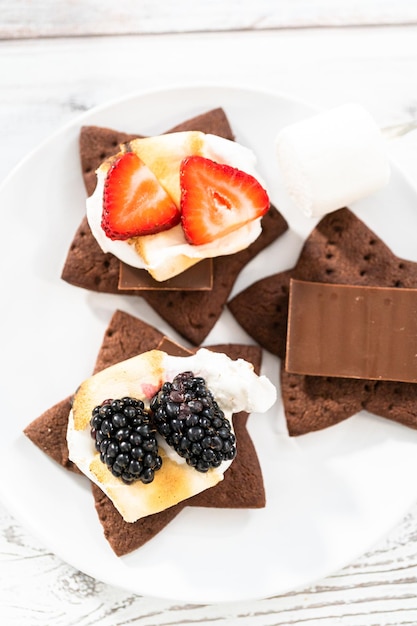 Photo making s'mores on a homemade star-shaped chocolate graham cracker with toasted marshmallow and fruits.