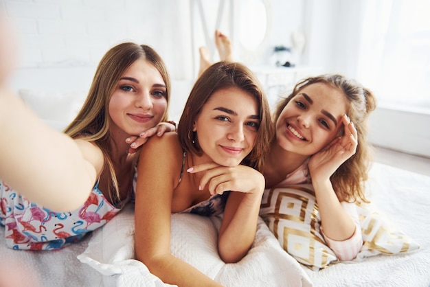 Making selfie. Happy female friends having good time at pajama party in the bedroom.