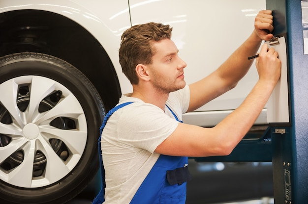 Making some notes about car. Handsome young man writing something in clipboard while standing in workshop with car in the background