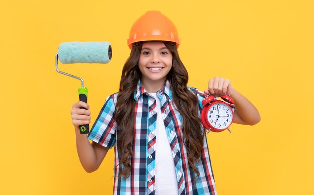 Making surface repair happy teen girl hold paint roller and alarm clock child in helmet