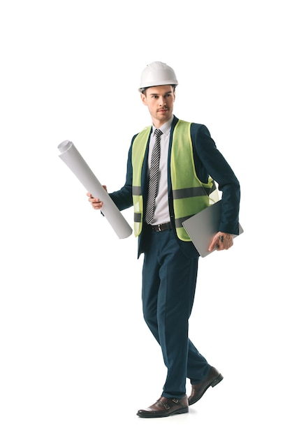 Male architect in hardhat and safety vest walking with blueprint and laptop isolated on white