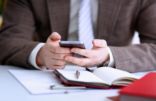 Male arm in suit hold phone and silver pen at workplace closeup Read news mania send sms chat addict use electronic bank modern lifestyle job plan colleague share blog tweet web application search