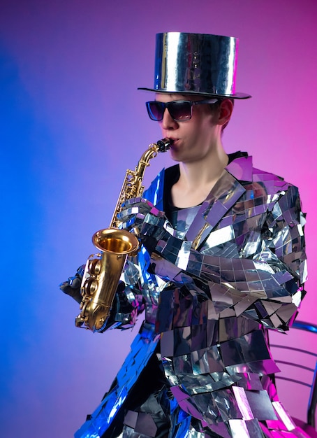 The male artist in a mirrored suit and a hat with a saxophone in neon light