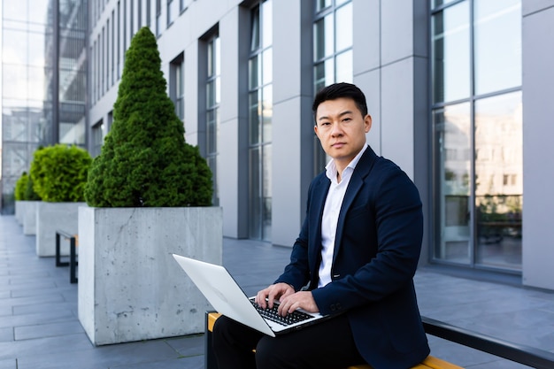 Фото Мужской азиатский бизнесмен, работающий на ноутбуке онлайн-банкинг на компьютере возле офисного центра, глядя в камеру