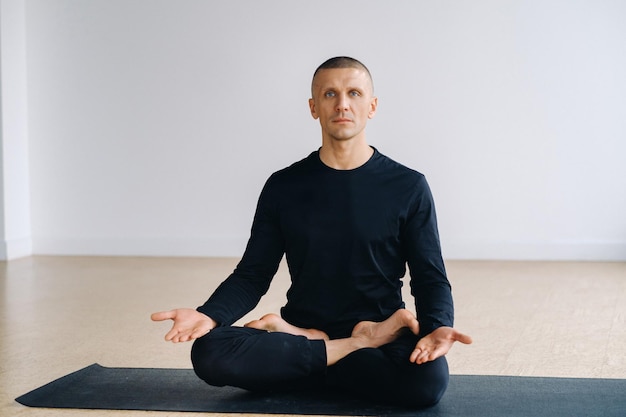 A male athlete in black sportswear is sitting on a mat in the lotus position in the gym