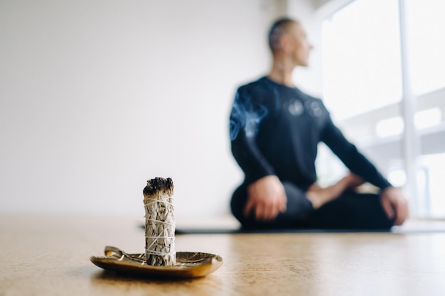 A male athlete in black sportswear sits in the lotus position against the background of an incense candle in the gym