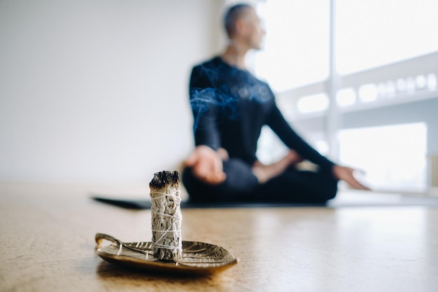 A male athlete in black sportswear sits in the lotus position against the background of an incense candle in the gym