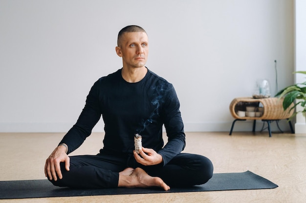 A male athlete in black sportswear sits in the lotus position in his hands with an incense candle in the gym