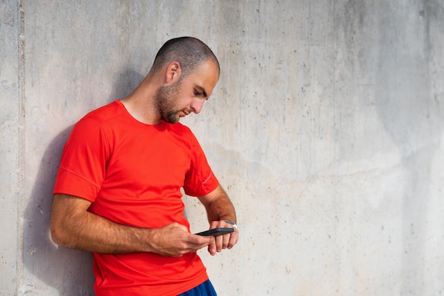 Photo male athlete synchronising data from sports watch with phone caucasian man in red sportswear concrete wall background spain alicante