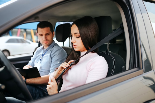 Male auto instructor takes exam with a  young woman. Brunette hold hands on seat belt and lock it. Young man sit besides with exam papers.
