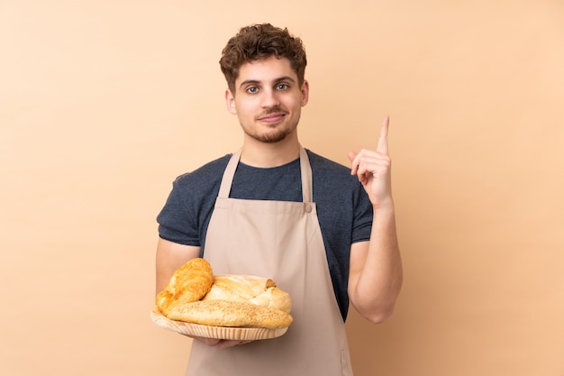 Male baker holding a table with several breads on beige pointing with the index finger a great idea