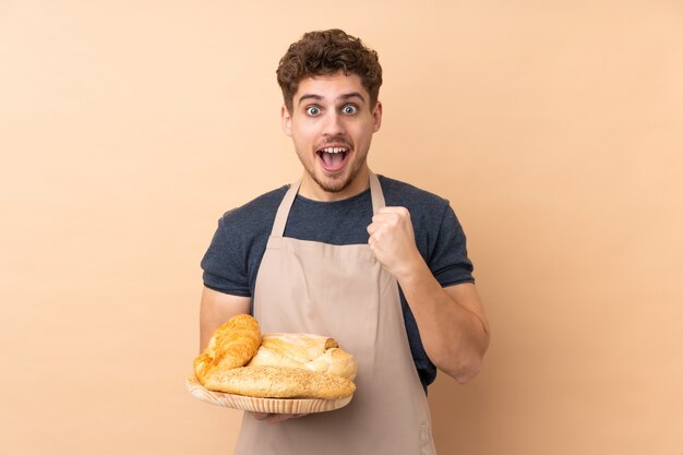 Male baker holding a table with several breads on beige wall celebrating a victory