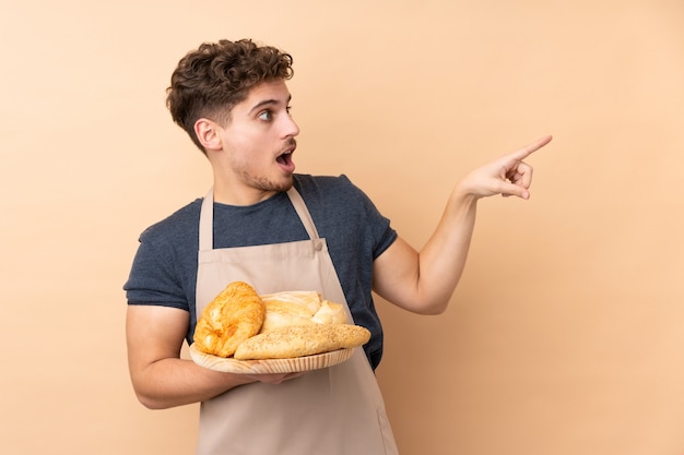 Male baker holding a table with several breads on beige wall pointing finger to the side