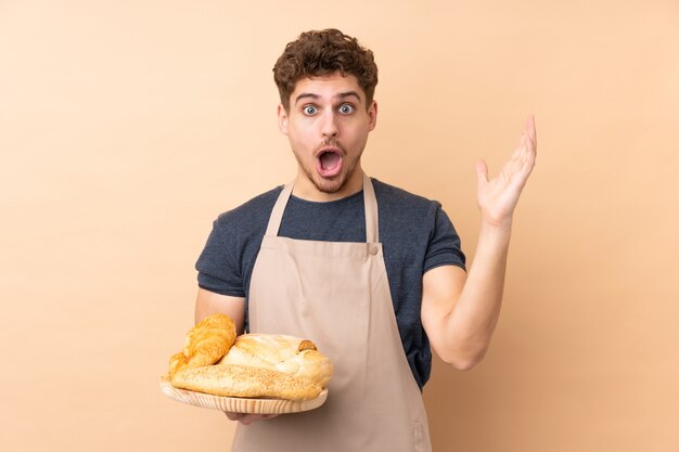 Male baker holding a table with several breads on beige with surprise facial expression