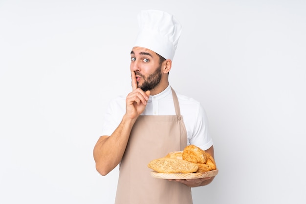 Male baker holding a table with several breads isolated on white doing silence gesture