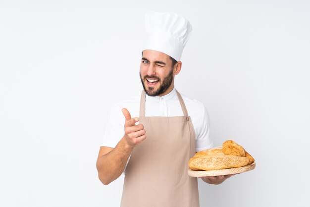 Male baker holding a table with several breads isolated on white wall points finger at you