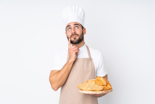 Il panettiere maschio che tiene una tabella con parecchi pani isolati sulla parete bianca che pensa un'idea