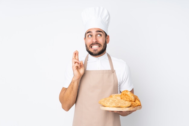 Male baker holding a table with several breads on white wall with fingers crossing