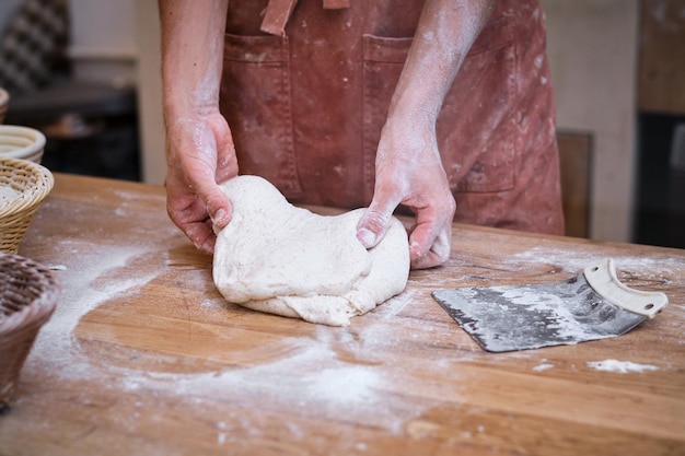 Male baker's hands working bread dough with flour on a wooden table.