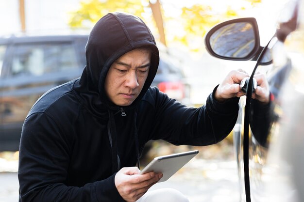 Maschio bandito ladro ladro d'auto asiatico utilizza un tablet per spegnere l'allarme dell'auto