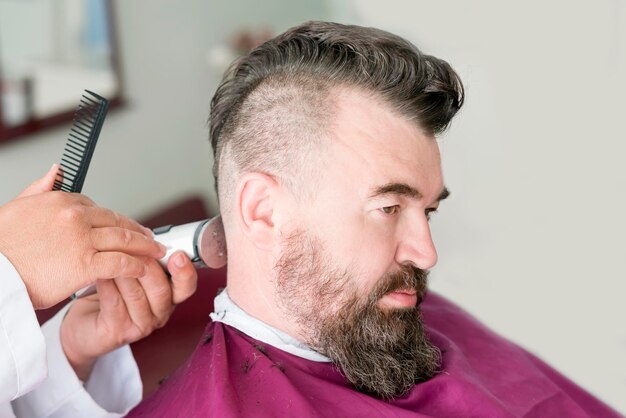 Male barber makes a mohawk hairstyle using clipper of a adult man with beard