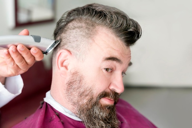 Мужской парикмахер делает прическу ирокезом с помощью машинки для стрижки волос взрослого мужчины с бородой