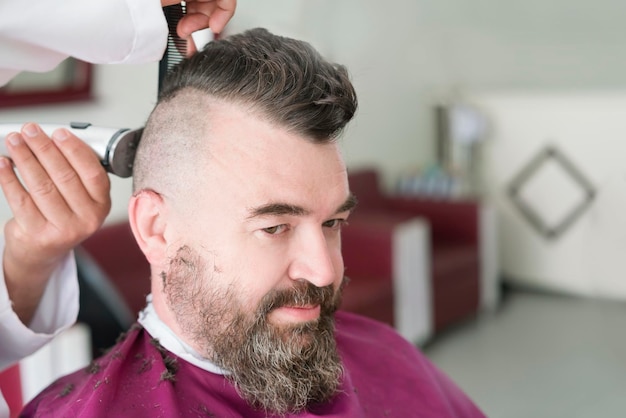 Male barber makes a mohawk hairstyle using clipper of a adult man with beard