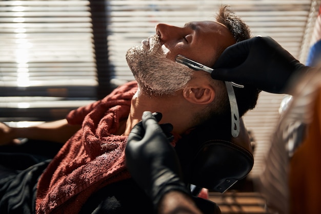 Фото Мужской парикмахер бреет мужскую бороду винтажной опасной бритвой