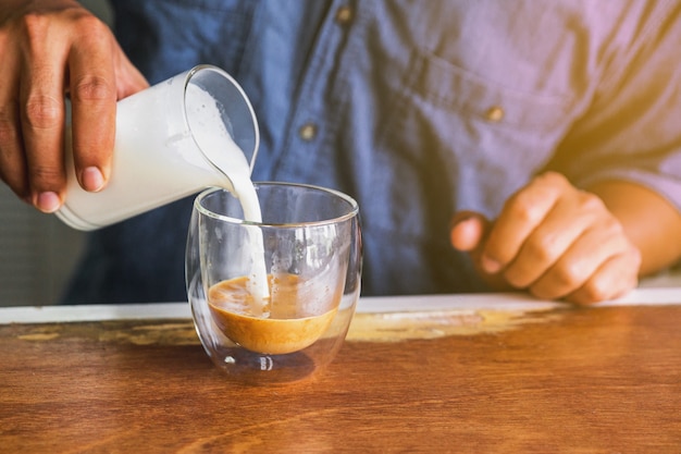 Male Barista pouring fresh milk in black coffee for making a latte at the wooden counter bar.