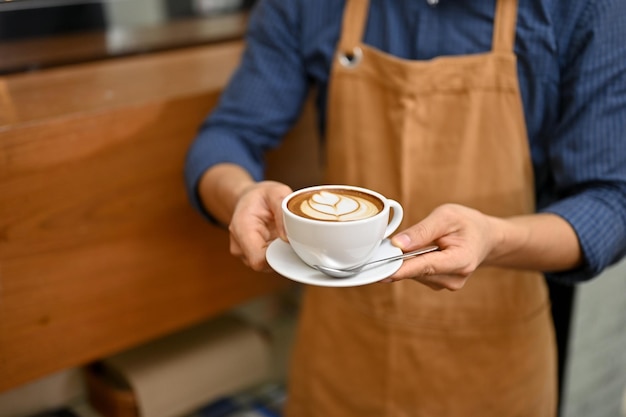 カフェのカウンターの前で美しいラテ アートのカップを保持している男性のバリスタまたはウェイター