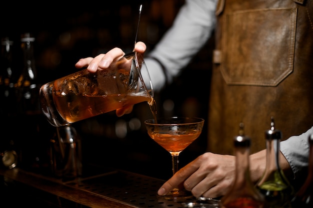 Male bartender pouring a brown alcoholic drink from the measuring cup to the cocktail glass