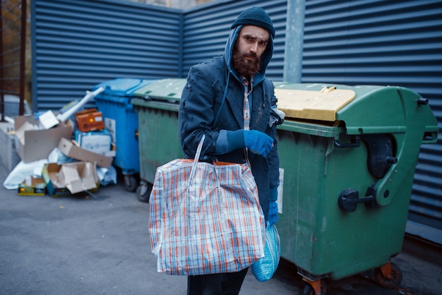 Male bearded beggar searching food in trashcan on city street.