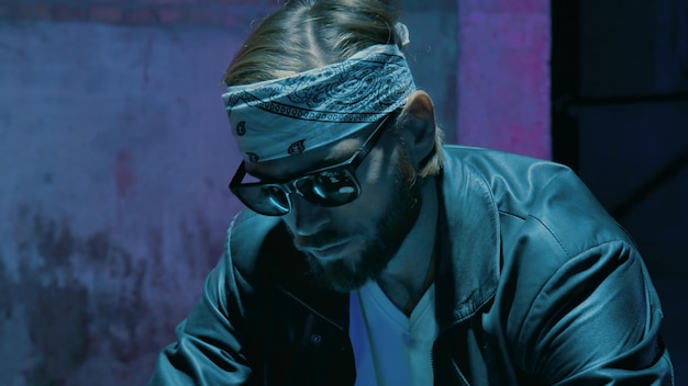 Male biker in a bandana in sunglasses against a brick wall with a neon sign