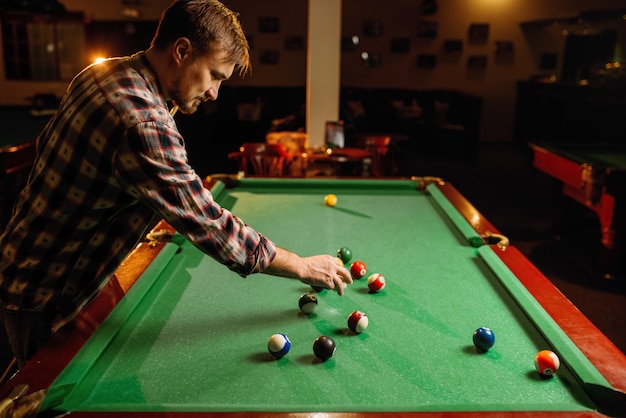 Male billiard player places balls on green table, poolroom interior. Man plays american pool game in sport bar